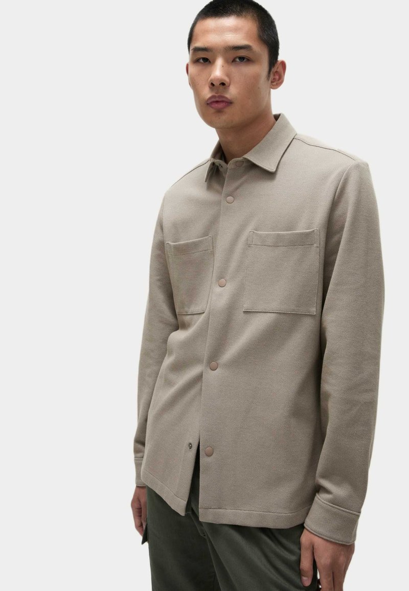 Chemise beige clair à manches longues avec une fermeture à boutons, deux poches poitrine et une texture lisse. Elle présente un col et des poignets à boutons.