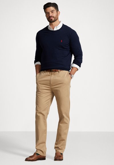 Jersey de marinero sobre una camisa con cuello, chinos color caqui y mocasines marrones. Cuenta con un pequeño logotipo rojo en el jersey. Texturas suaves en todo.