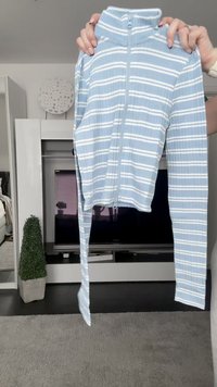 Lyseblå zip-up jakke med hvite striper, lange ermer og høy krage. Stoffet ser glatt ut med en lett tekstur.
