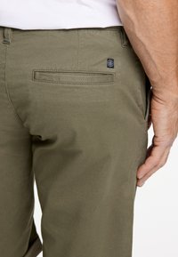Pantaloni verde oliva con tessuto strutturato, dotati di una tasca posteriore e un piccolo patch con logo. Vestibilità sartoriale con passanti per cintura e chiusura con zip.