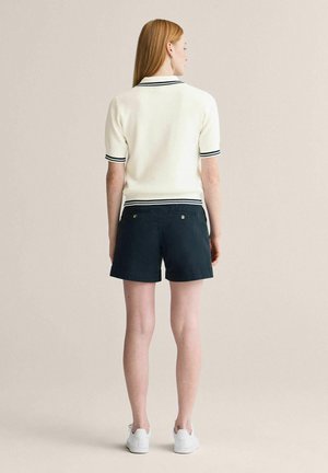 Suorahiuksinen punatukkainen nainen kuvattuna takaa, valkoisessa lyhythihaisessa polopaidassa mustilla reunoilla, tummansinissä shortseissa ja valkoisissa tennareissa, seisoo beigenväristä taustaa vasten.