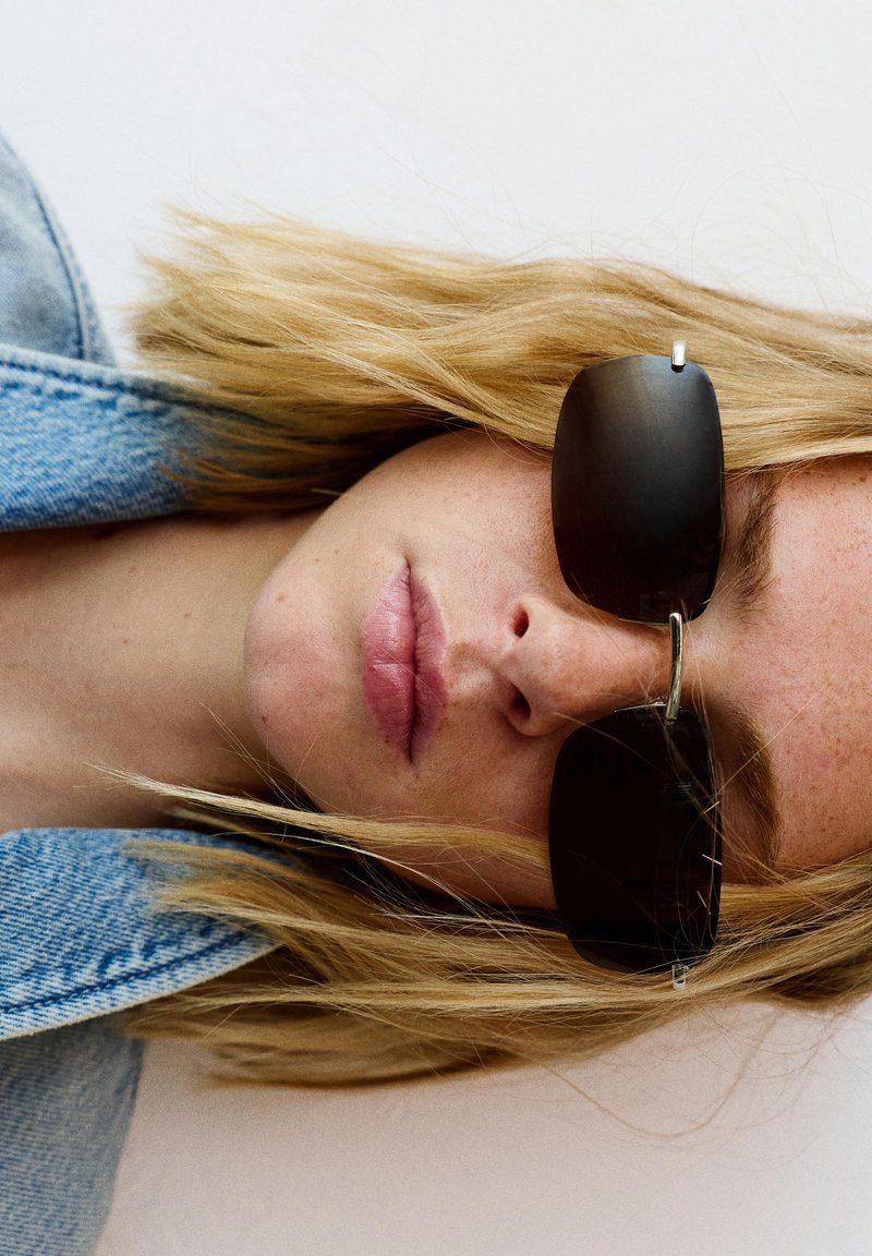 Femme aux longs cheveux blonds porte des lunettes de soleil noires rectangulaires et une veste en jean, vue en gros plan en angle.