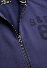Sudadera azul con cremallera y la inscripción "S & D 6" en el pecho, cremallera plateada con la etiqueta "Super Dry" y etiqueta interior de tela visible.
