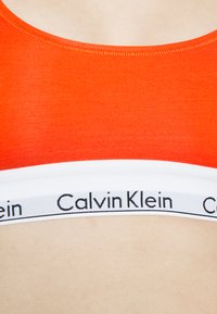 Piros sportmelltartó sima anyaggal, kerek kivágással és a "Calvin Klein" logóval ellátott fehér pánttal. Puha tapintású, minimális varrási részletekkel.