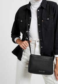 Černá kabelka přes rameno s texturovaným povrchem, obdélníkovým tvarem a tenkým nastavitelným popruhem. Na přední straně se nachází nenápadný detail s logem.