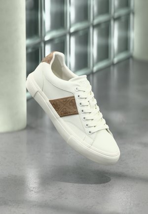 Witte lage sneaker met beige patroon aan de zijkant en hielpatch, witte veters, zwevend boven een grijze vloer in een moderne binnenomgeving.