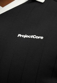 Svart polo-shirt med vit krage och vertikala ränder. Har "ProjectCore"-logotypen i vitt, centralt placerad på bröstet.