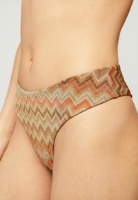 Primer plano de una persona que lleva braguitas de bikini de corte alto con un patrón de zigzag multicolor en tonos tierra.