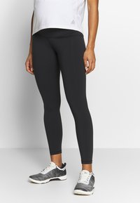 Leggings pretos feitos de tecido elástico, com uma cintura alta. Combinados com sapatilhas atléticas cinzas e brancas, num fundo simples.