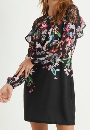Femme portant une robe noire à fleurs avec des manches longues transparentes et des détails en dentelle aux épaules, ajustant une manche avec ses mains.