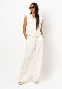 Witte mouwloze cropped top met gestructureerde strepen, gecombineerd met losse, bijpassende high-waisted broek. Zilveren puntige hakken en zonnebril.