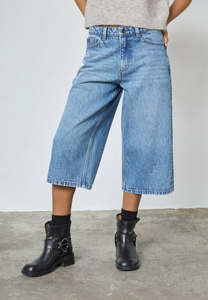 Person iført højtaljede, lyseblå cropped bukser med vide ben, sorte ankelstøvler og sorte sokker, stående på betongulv.