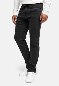 Svart denimjeans med slim fit. Har femlommedesign, subtil søm, og er kombinert med hvite joggesko og en langermet skjorte.