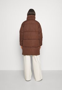 Hnědá péřová bunda s vysokým límcem a horizontálním prošíváním, sladěná s krémovými širokými kalhotami. Bunda má hladký, matný povrch.