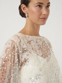 Γυναίκα με καστανά μαλλιά σε κότσο, φορώντας διάφανο λευκό μπλουζάκι διακοσμημένο με υφασμάτινα λουλούδια, πέρλες και ασημένιες παγιέτες, στραμμένη προς τα δεξιά.