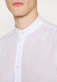 Camisa de lino blanca con cuello mao, que presenta un tapeta de botones y una textura suave. La superficie ligeramente arrugada añade un detalle sutil.