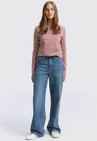 Pruhovaná košeľa s dlhým rukávom v jemnej ružovej a bielej farbe, doplnená o džínsové modré široké nohavice s vysokým pásom. Outfit dopĺňajú biele tenisky.