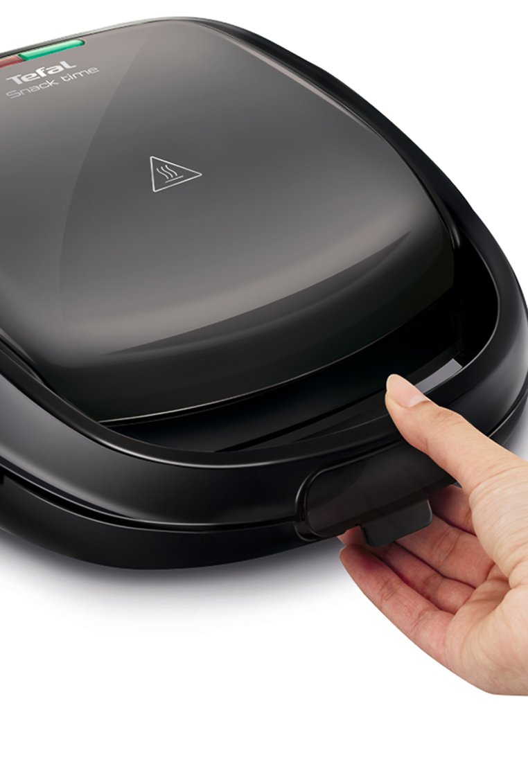 Aparato Tefal Snack time, de plástico negro, forma redondeada, tapa abierta mostrando las placas de parrilla, mano agarrando el cierre. Luces indicadoras en verde y rojo.