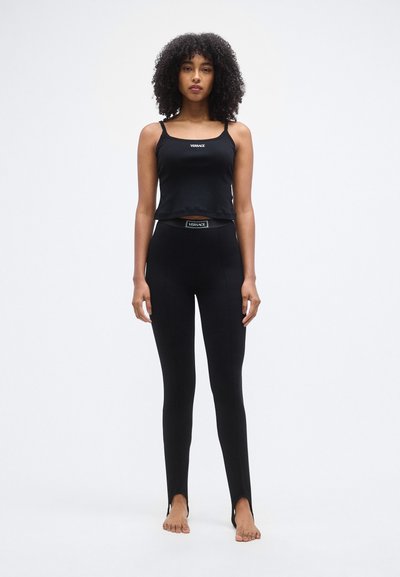 Haut court noir à fines bretelles et logo, associé à des leggings noirs taille haute avec des fentes à la cheville. Texture de tissu lisse.