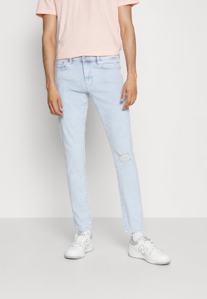 Personne portant un jean déchiré bleu clair, une chemise rose pâle à manches courtes, des chaussettes blanches et des baskets blanches New Balance, debout devant un fond uni.