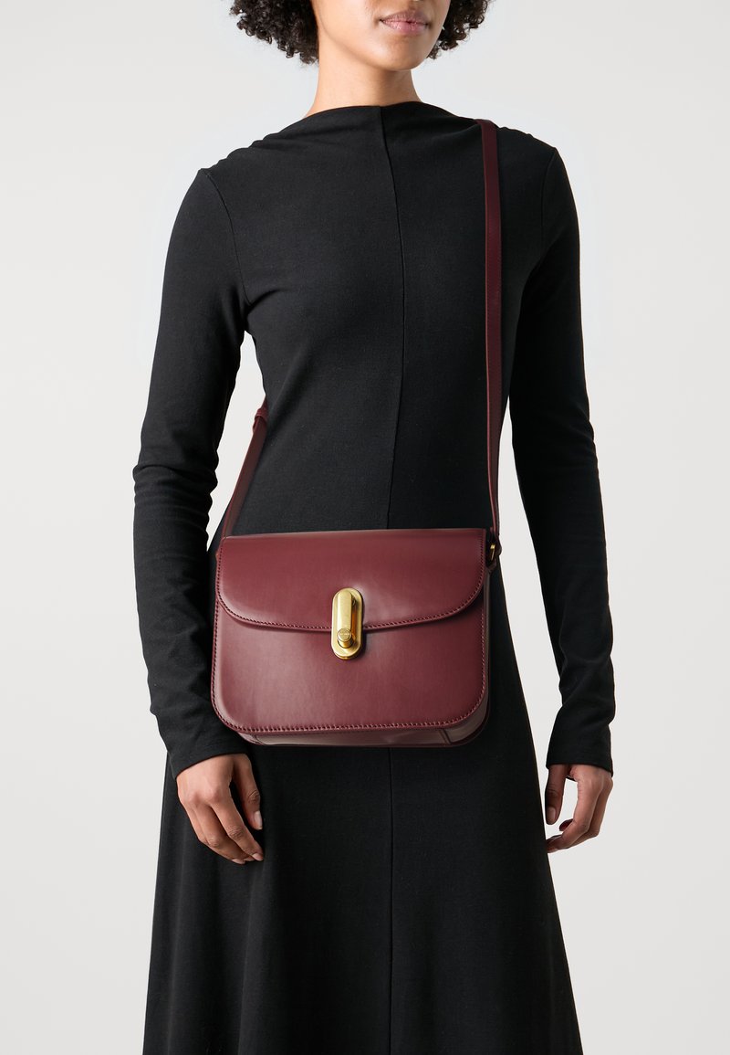 Burgundská kožená crossbody taška so zapínaním na klopu, zlatými doplnkami a hladkou textúrou. Ramenný popruh je nastaviteľný a má elegantný dizajn.