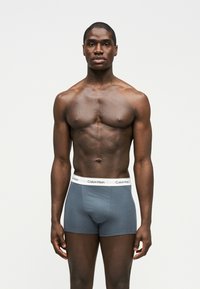 Hombre musculoso de pie de frente, vistiendo calzoncillos bóxer grises de Calvin Klein con una cintura blanca, contra un fondo claro y liso.