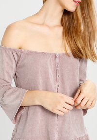Off-the-shoulder, licht mauve blouse met klokmouwen, knopen aan de voorkant en een licht textuurstof. Gladde afwerking met een gekruide zoomdetail.