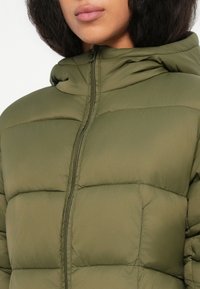 Mujer vistiendo una chaqueta acolchada verde oliva con capucha, mostrando la parte superior del torso y parte de la cara sin que se vean los ojos.