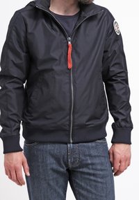 Svart zip-up jacka med en något glänsande yta, elastiska ärmar, röd dragkedjedragare och ett runt logotypmärke på vänster ärm.