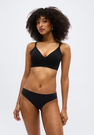 Γυναίκα με σγουρά μαλλιά που φοράει μαύρο σουτιέν και ταιριαστό μαύρο εσώρουχο, στέκεται μπροστά σε απλό ανοιχτόχρωμο φόντο.