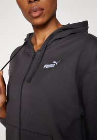 Felpa nera con zip realizzata in tessuto morbido, dotata di cappuccio regolabile con lacci e logo Puma viola sul petto. Ideale per un look casual.
