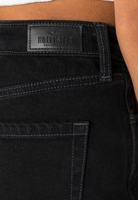 Jeans pretos em denim com um acabamento texturizado, apresentando uma etiqueta em couro com a inscrição "Hollister California" e detalhes de costura cinza em contraste.