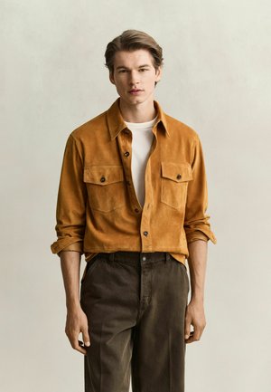 Young man wearing a mustard buttoned shirt over a white T-shirt and dark pants, standing with arms relaxed against a plain background.