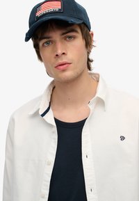 Gorra azul marino con un parche de la bandera de EE. UU., camisa blanca de botones y camiseta oscura de color marino debajo; tejido suave sin patrones prominentes.