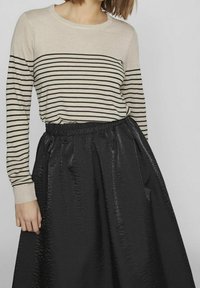 Model w paskowanej beżowo-czarnej koszuli z długim rękawem, zestawionej z czarną, błyszczącą spódnicą w falbany, na neutralnym tle.