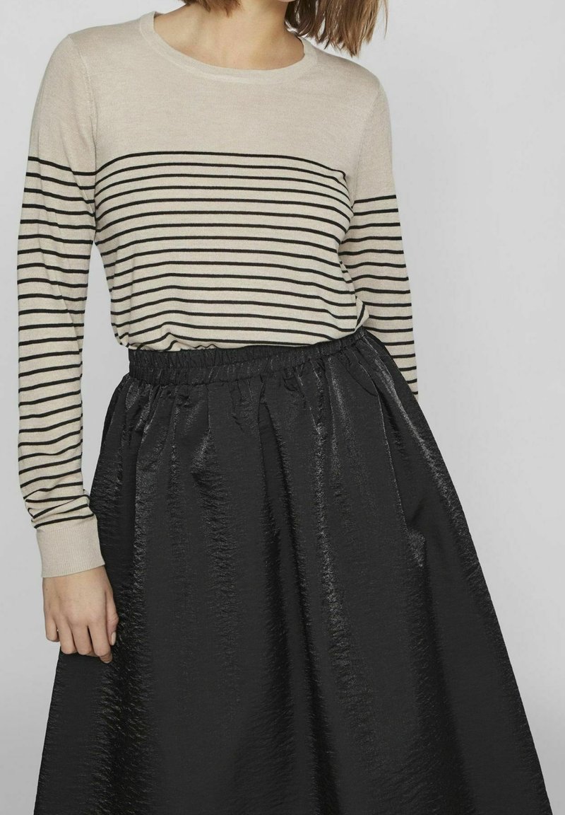 Model w paskowanej beżowo-czarnej koszuli z długim rękawem, zestawionej z czarną, błyszczącą spódnicą w falbany, na neutralnym tle.
