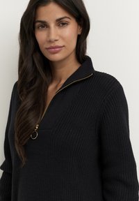 Pull en tricot noir avec un col zippé quart, doté d'une tirette dorée et d'un tissu texturé. Pas de motifs ni d'accents supplémentaires visibles.