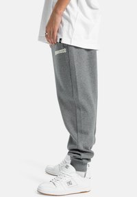 Graue Jogginghosen mit gerippten Bündchen und seitlichen Streifen, mit einem weißen gestickten Logo. Kombiniert mit weißen Sneakers.