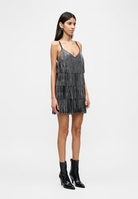 Μαύρο φόρεμα με φράντζα και V-λαιμόκοψη, με στρώσεις από λωρίδες και λεπτούς ιμάντες ώμων. Φορεμένο με μαύρα μποτάκια αστραγάλου.