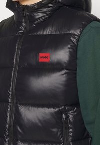 Schwarze gesteppte Weste mit Reißverschluss und Kapuze. Verfügt über ein rotes, rechteckiges Logo-Patch. Darunter ist ein dunkelgrünes Langarmshirt sichtbar.