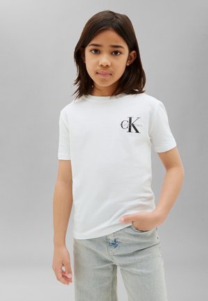 Młoda osoba o ciemnych włosach do ramion, ubrana w białą koszulkę Calvin Klein i jasnoniebieskie dżinsy, stojąca na jednolitym szarym tle.