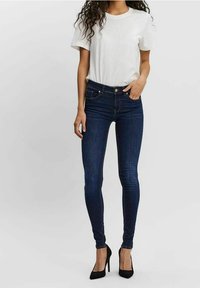 Jean skinny bleu foncé associé à un t-shirt blanc uni, porté avec des escarpins noirs. Doté de cinq poches et d'une coupe ajustée.