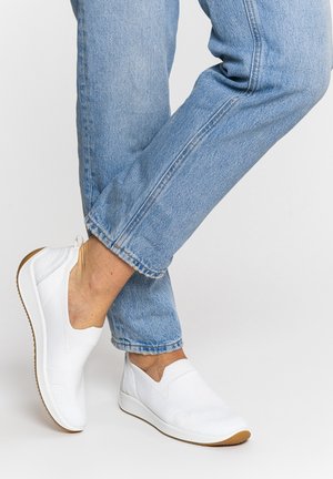 Person trägt hellblaue Jeans und weiße Slip-On-Sneaker und steht mit einem Bein über das andere geschlagen vor einem schlichten Hintergrund.