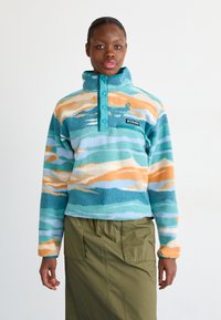 Pullover de fleece com um colarinho alto, apresentando padrões de ondas em azul-turquesa, laranja e azul, quatro botões azul-turquesa e um pequeno logo preto.