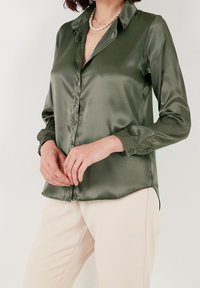 Green satin button-up shirt with a collar, long sleeves, and a curved hem; worn with beige pants and layered with a pearl necklace.