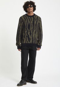 Pull en tricot olive avec des rayures abstraites bleu marine, doté d'un col rond, de poignets et d'un ourlet côtelés; associé à un pantalon et des chaussures noirs.