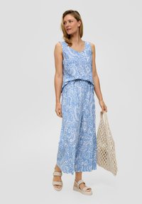 Jasnoniebieski jumpsuit bez rękawów z białymi kwiatowymi wzorami, szerokimi nogawkami i zebranym w pasie. Model trzyma plecioną beżową torbę.