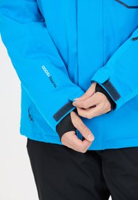 Blå vattentät jacka med en slät yta, med kardborrefästen vid ärmarna och en logotyp på ärmen. Händer som justerar ärmfästet syns.