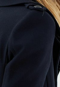 Námořnický modrý vlněný kabát s konstrukčně tvarovaným límcem, texturovanou tkaninou a detailním knoflíkem na rameni pro zvýraznění.
