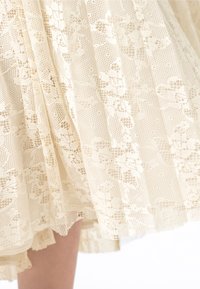 Spetsklänning i ljus krämfärgad med intrikata blomsterdesigner, plisserad stil och en texturerad yta. Nederdelen har en vågig kant.